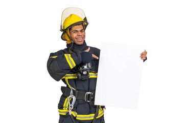 Young smiling African American fireman wearing uniform and helmet looking to the camera and holding blank paper sheet in hand and finger pointing. Copy space for text, isolated on white background 