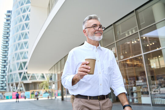 Happy Older Senior Professional Business Man, Smiling Gray Haired Mature 60 Years Old Businessman Wearing White Shirt Going Outdoors Holding Coffee Walking In The Big City.