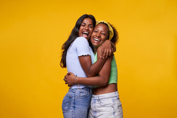 Two diversity pretty positive girls toothy smile hugging isolated on yellow color background. Young lesbian lovers couple embracing and looking camera. Friendship and homosexual relationship. - obrazy, fototapety, plakaty