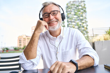 Happy older senior business man wearing headphones looking at webcam having hybrid remote video...
