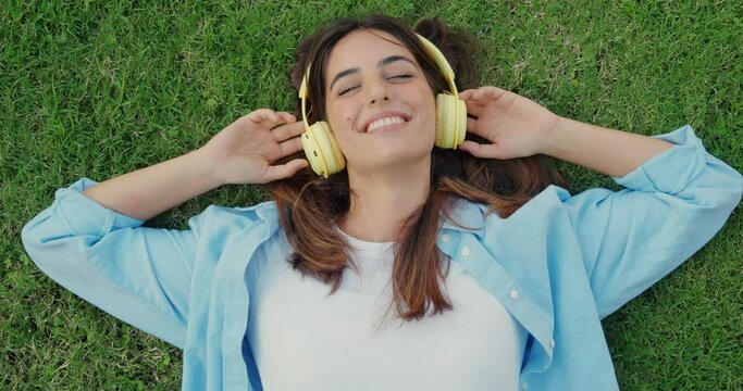  Young cheerful millennial woman lying on the grass listening to music with headphones