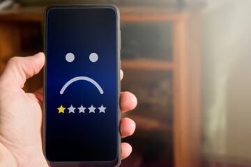 Closeup of male hand holding mobile phone with sad face and one star rating. Very unhappy customer...