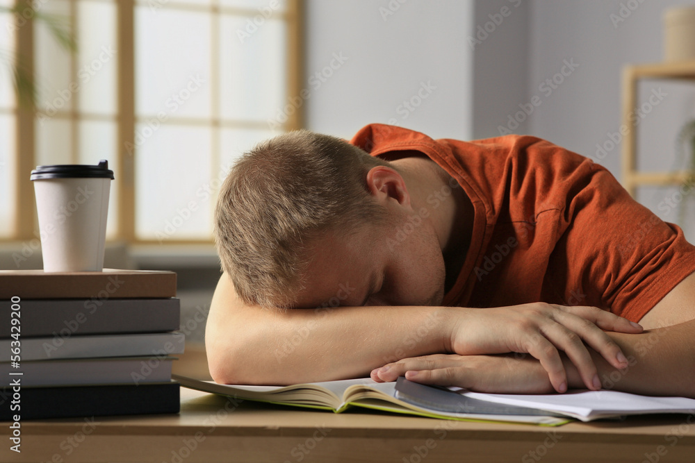 Wall mural Tired man sleeping near books at wooden table indoors