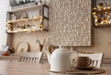 White teapot with cup on wooden table in kitchen, space for text