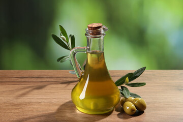Jug of cooking oil, olives and green leaves on wooden table against blurred background