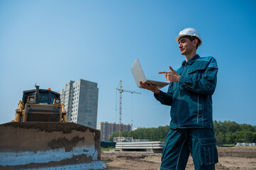 A male builder in a helmet stands against the background of a construction site with a laptop in his hands. 