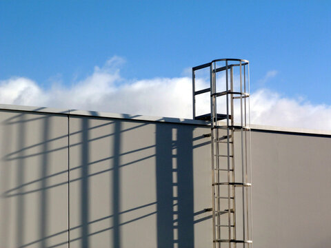 aluminum roof access ladder with safety cage. elevation view. strong shadows on white concrete wall. rungs and vertical safety bars. buildings, architecture and technology. construction. blue sky 