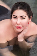 Portrait of brunette xxl young woman in black swimsuit relaxation lying down in outdoor pool at balneotherapy spa, hot springs resort. Authentic outsize young woman looking at camera. High angle shot - obrazy, fototapety, plakaty