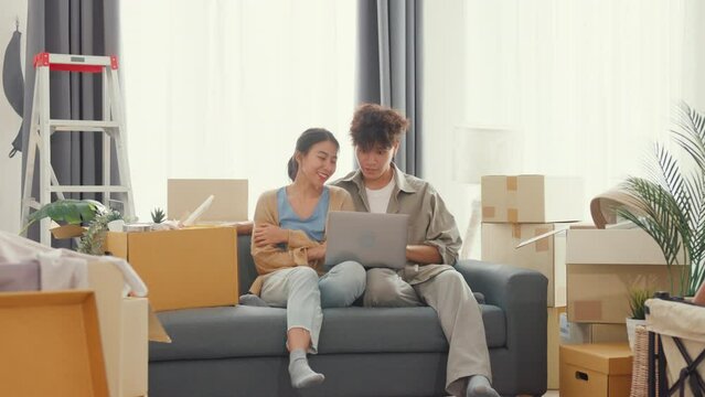 Young happy Asia couple sit on sofa couch use computer laptop online shopping furniture from decoration store in living room at home. Husband wife move new house, Apartment Rent And Ownership Concept.