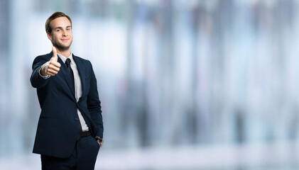 Smiling businessman showing thumb up like sign gesture, in black suit, over blurred interior...