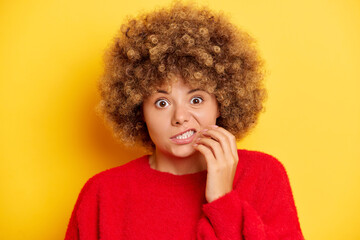 Worried woman looks nervously, has curly hair, bites finger nails with scare look, anticipates what...