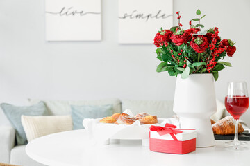 Vase with flowers, pastries, glasses of wine and gift for Valentine's Day on dining table in room