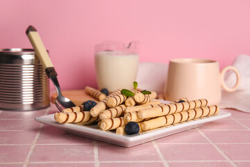Plate with tasty wafer rolls, blueberries, glass of milk and tin can on pink table