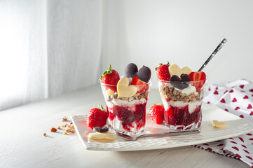 Sweet breakfast on Valentine's Day on a white wooden table. Creamy dessert with ripe strawberries and pureed raspberries, muesli in a glass decorated with chocolate in the shape of a heart. Side view