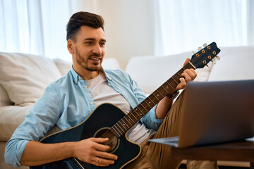 Happy man plays acoustic guitar while surfing the net on laptop at home.