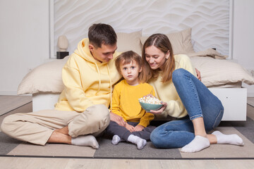 Friendly family, lovely parents in yellow clothes with child son sitting by the bed and eating popcorn