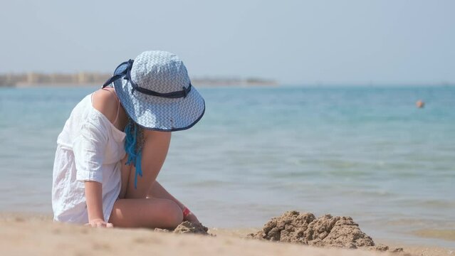Happy hild girl in big hat and white dress playing alone with wet sand on sandy beach near clear sea lagoon water