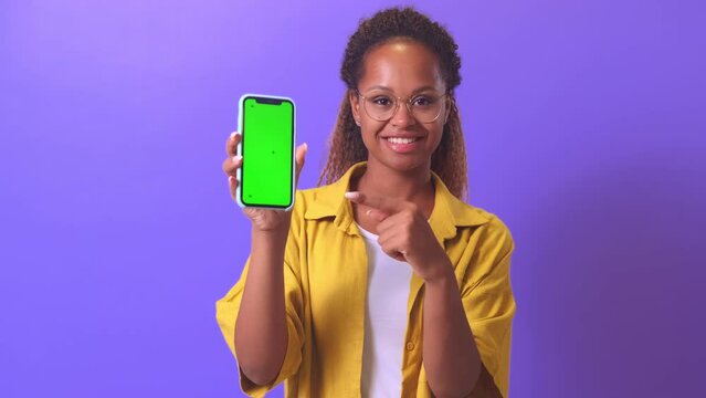Young cheerful African American woman holds phone pointing with finger at green display to recommend downloading cool youth application or registering on interesting site stands in lilac studio.