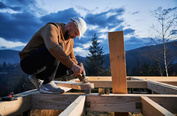 Man worker building wooden frame house on pile foundation. Carpenter installing furniture for wooden joist, using hammer and screwdriver. Carpentry concept.