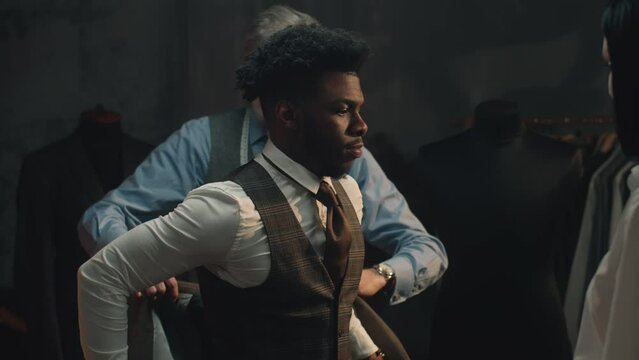 African American young businessman on suit fitting in stylish luxury designer atelier. Tailors put jacket on man. Tailored wedding or business suit. Concept of fashion, handmade and couturier.
