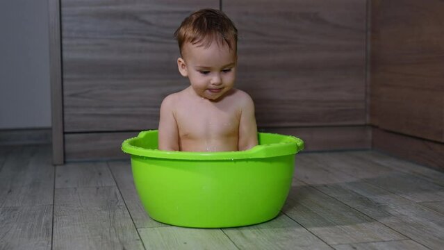 Happy Caucasian baby splashing water out of washtub he’s sitting in. Naked cute child making faces, showing tongue to camera.