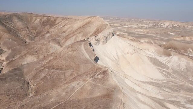 Aerial drone flying towards desert mountain with sharp ridges with trail going downhill, during sunny bright day with blue sky, near Dead Sea, Arad Mountains, Negev desert, Israel