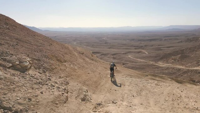 Aerial Mountain biking downhill alone in the desert in Mizpe Ramon, Negev, Israel, during a sunny day with blue skies drone follow me active track