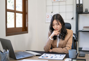 Woman sitting at table doing planning financial report, business plan investment, finance analysis concept.Young thoughtful asian business woman wearing suit working in office, analyzing document