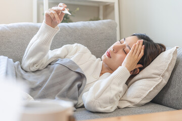 Sick, influenza asian young woman, girl lying sore on sofa, have a fever, hand holding thermometer for check measure body temperature, illness while sitting rest on sofa at home. Health care concept.