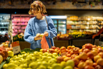 Young woman chooses apples to buy and puts them into mesh bag at fruit department of a supermarket. Concept of consumerism of healthy food and use of reusable bags