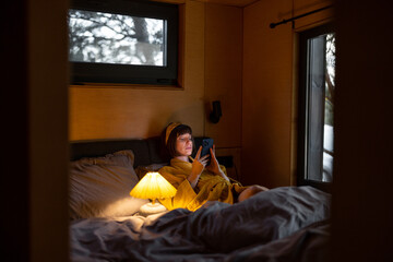 Naklejka premium Woman in yellow bathrobe uses a smartphone while lying in bed at tiny bedroom of wooden cabin on nature. Communication online during bedtime and addiction to smartphone