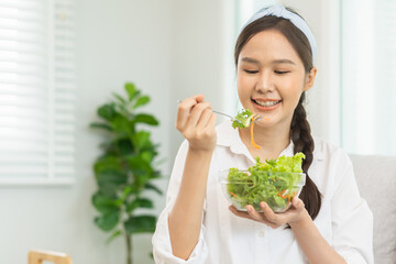 Diet, Dieting asian young woman, girl hand use fork at lettuce on mix vegetables, green salad bowl, eat food low fat to good body sitting on sofa at home. Nutritionist female, Weight loss for healthy.