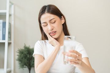 Suffering from toothache, blur asian young woman touch cheek, face expression ache or feel pain, sensitive molar teeth, hand holding glass of water with ice, inflammation when drink cold, healthcare.