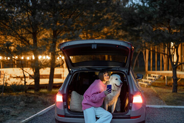 Portrait of a young woman with her cute white dog sitting together at car trunk, traveling on nature during the evening time