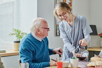 Young caregiver pouring coffee from teapot for senior man while he having breakfast at table - Powered by Adobe