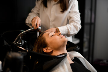 handsome female client relaxed lying on hair washing chair with her eyes closed while hairdresser...