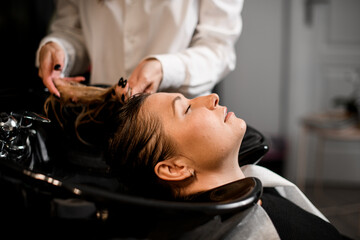 attractive female client relaxed lying on hair washing chair with her eyes closed while hairdresser washes her hair