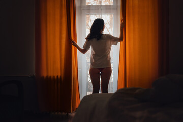 New day. Silhouette of woman opening orange curtains in morning at window. Rear view. Concept of freedom, hope and happiness