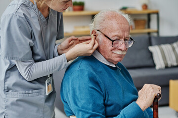 Young caregiver helping to wear hearing aid to senior man while she caring about senior people in...