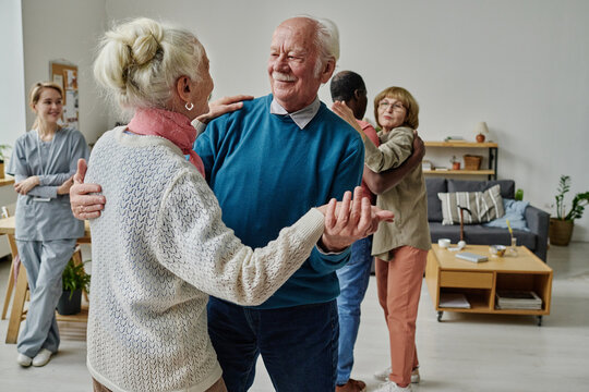 Happy Senior Couple Dancing Together In The Room With Other People And Caregiver In Background