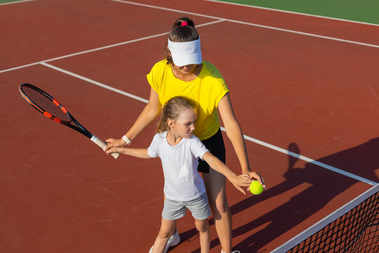 Cheerful coach in sports clothing teaching child to play tennis while both standing on tennis court.