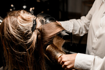 Stylist puts female hair with dryer and round comb in hairdresser. Close-up