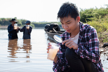 Young asian boy holds transparent tube which has example water inside to do the experiment and ph level measurement as his school project work with his friends behind at the river where he lived.