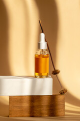 Serum is orange in transparent glass bottle with white dropper lid on wooden stand with branch of deadwood. Mockup of essential oil for care of women's skin on brown background, in rays of sunlight