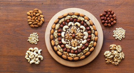 Obraz na płótnie Canvas Beautiful homemade festive chocolate cake decorated with various types of nuts on wooden table. Walnuts, hazelnuts, cashews, pine nuts and almonds next to it. Flat lay, close-up, top view, mock up