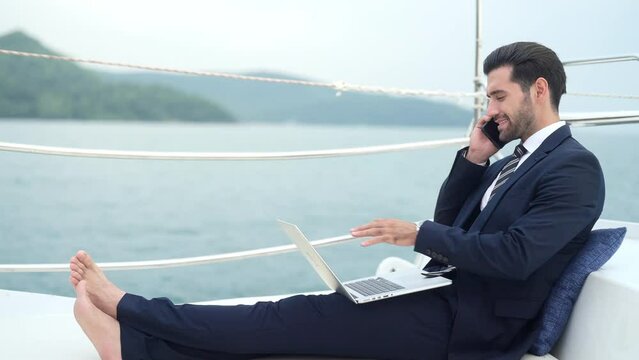 Businessman working on laptop computer and talking on mobile phone for global corporate business while travel on luxury private catamaran boat yacht sailing in the ocean on summer vacation at sunset.