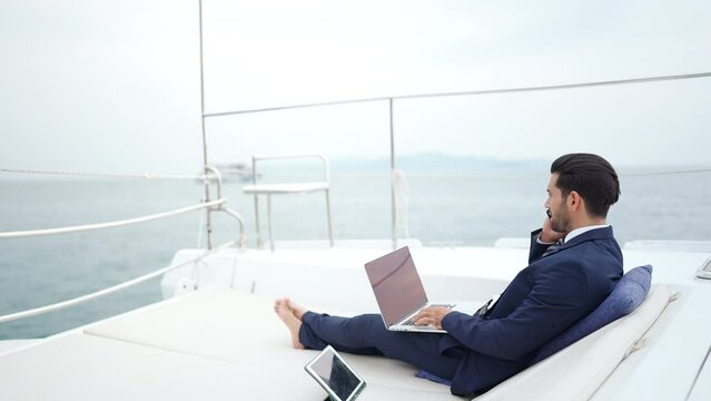 Businessman working on laptop computer and talking on mobile phone for global corporate business while travel on luxury private catamaran boat yacht sailing in the ocean on summer vacation at sunset.