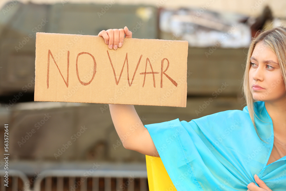 Wall mural sad woman wrapped in ukrainian flag holding poster with words no war outdoors