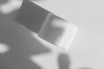 Face cream in white jar on light background with shadows from leaves. Mockup of clean package for...