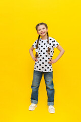 A little girl with beautiful long pigtails in jeans on a yellow isolated background, holds her hands on her hips and smiles.